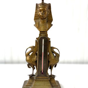 Op de afbeelding: Antieke messing thermometer met een Egyptische farao buste. De thermometer heeft een rode vloeistofkolom en wordt geflankeerd door twee vogelsculpturen. De basis is rechthoekig met decoratieve details.