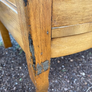 Puede incluir: Primer plano de una c&oacute;moda de madera con un acabado descascarado y desgastado. La c&oacute;moda tiene un frente curvo y un solo caj&oacute;n. La madera es de color marr&oacute;n claro con vetas marr&oacute;n oscuro.