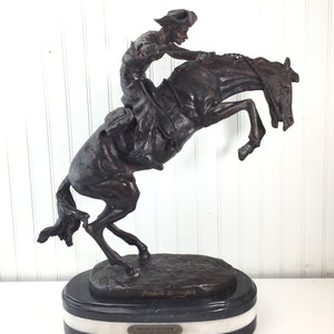 May include: A bronze sculpture of a cowboy riding a bucking bronco, on a black and white marble base. The sculpture is signed "Frederic Remington" and the base reads "Bronco Buster by Frederic Remington".