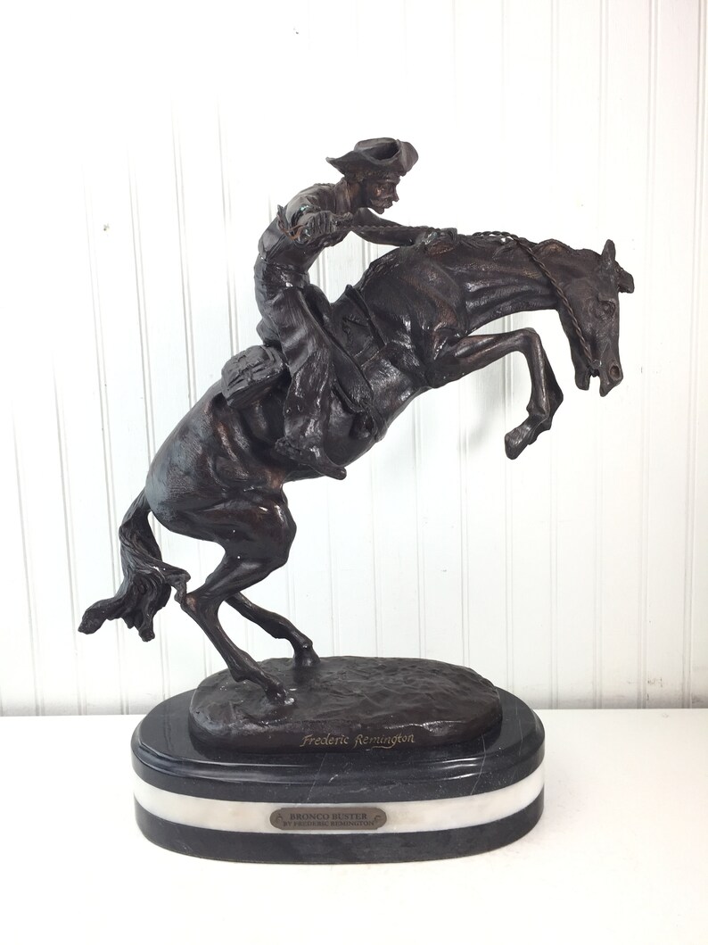 May include: A bronze sculpture of a cowboy riding a bucking bronco, on a black and white marble base. The sculpture is signed "Frederic Remington" and the base reads "Bronco Buster by Frederic Remington".
