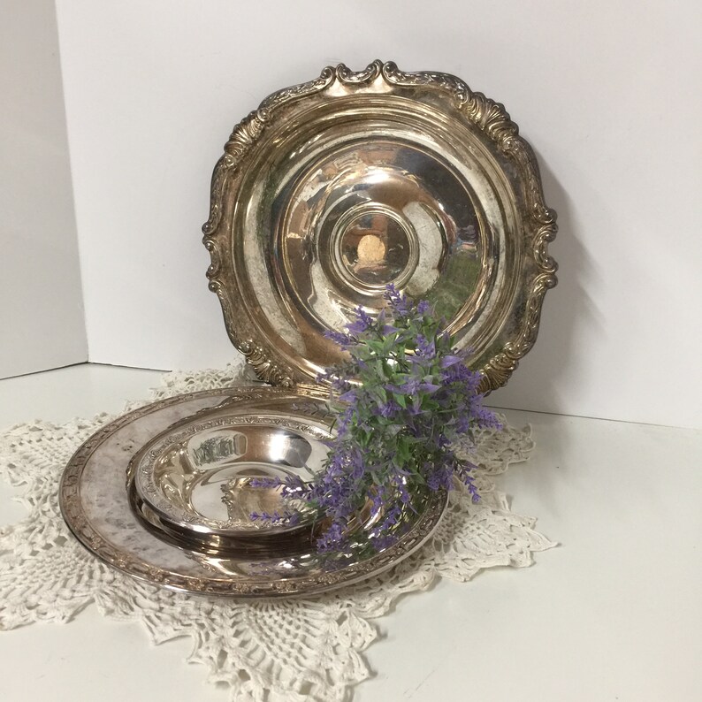 Puede incluir: Dos platos de servir plateados con bordes ornamentados. El plato m&aacute;s peque&ntilde;o est&aacute; anidado dentro del plato m&aacute;s grande. Un ramo de flores moradas descansa sobre los platos.