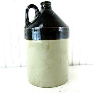 May include: A brown and white glazed stoneware crock with a handle. The crock has a dark brown top and a light brown bottom.