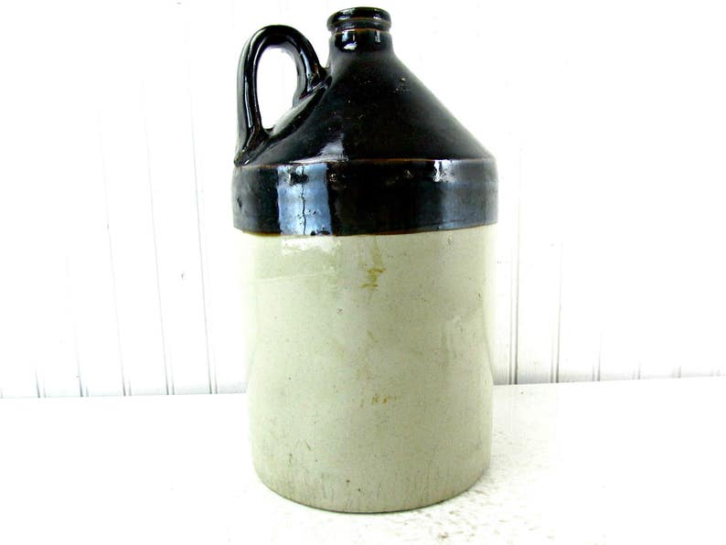 May include: A brown and white glazed stoneware crock with a handle. The crock has a dark brown top and a light brown bottom.
