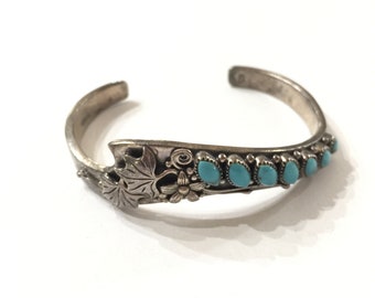 May include: A silver bracelet with a turquoise inlay design. The bracelet features a floral motif and a row of turquoise stones.