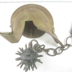 May include: A rusty metal helmet with a spiked ball attached to a chain. The helmet is open on the top and has a small, round opening on the side. The ball is covered in spikes and is connected to the helmet by a chain with a metal ring.