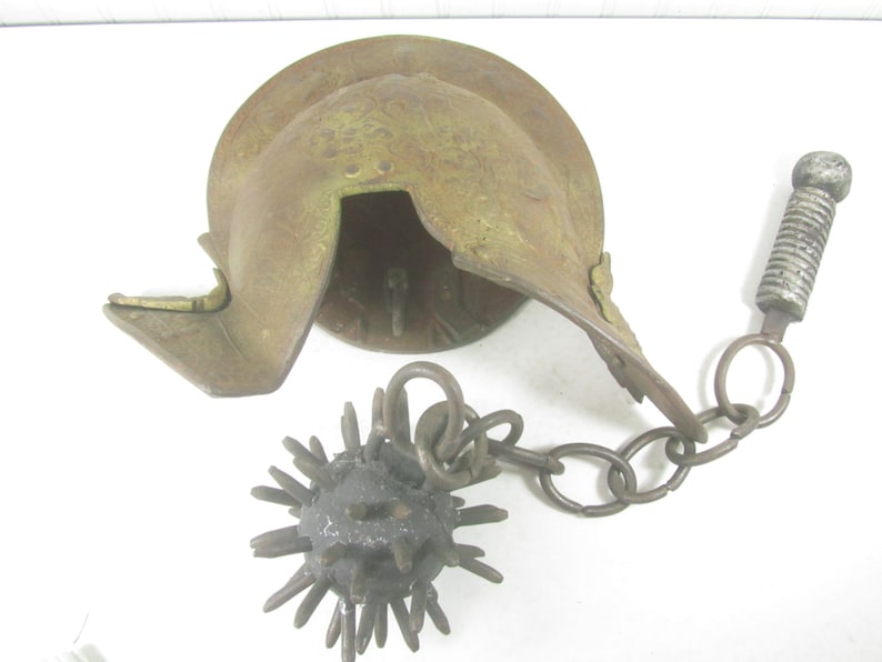 May include: A rusty metal helmet with a spiked ball attached to a chain. The helmet is open on the top and has a small, round opening on the side. The ball is covered in spikes and is connected to the helmet by a chain with a metal ring.