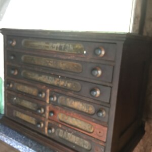 May include: A dark wooden cabinet with six drawers. Each drawer has a glass label with a faded inscription. The labels read "BUTTONS", "SEWING", "YARNS", "NEEDLES", "TRIMMING", and "DAMASK"