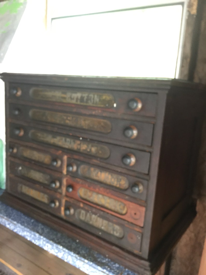 May include: A dark wooden cabinet with six drawers. Each drawer has a glass label with a faded inscription. The labels read "BUTTONS", "SEWING", "YARNS", "NEEDLES", "TRIMMING", and "DAMASK"