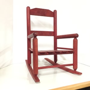 Puede incluir: Una silla mecedora de madera roja con un asiento de listones y un respaldo redondeado. La silla est&aacute; hecha de madera maciza y tiene un estilo r&uacute;stico, de granja.