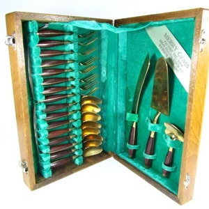 May include: A wooden box with a green velvet lining contains a set of gold-toned cutlery with brown handles. The box is labeled "Merry Gems" with the text "Factory 7244 Hong Kong" and "Made in Hong Kong".