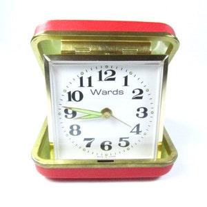 May include: A red, leather-covered, square-shaped alarm clock with a white face and gold-colored hands. The clock face has black numbers and the words "Wards" printed in black.