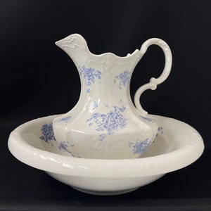 May include: White porcelain pitcher and washbasin set with blue floral patterns. The pitcher has a curved handle and a spout, and the washbasin is round with a wide rim. The set is likely vintage or antique.