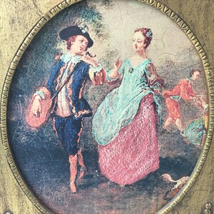 May include: An oval painting framed in gold, depicting a couple in a garden setting. The man is wearing a blue jacket and a black hat, while the woman is wearing a blue and red dress. The frame has ornate details.