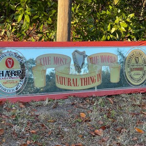 K&ouml;nnte beinhalten: Ein roter Holzrahmenspiegel mit einer reflektierenden Oberfl&auml;che. Der Spiegel zeigt eine Vintage-Werbung f&uuml;r Harp Lagerbier und Guinness Extra Stout. Der Text auf dem Spiegel lautet "Das nat&uuml;rlichste der Welt".