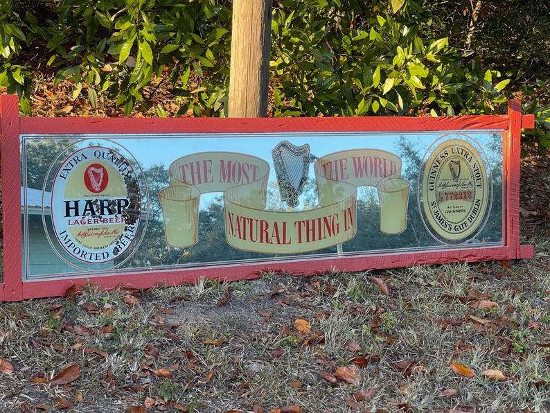 K&ouml;nnte beinhalten: Ein roter Holzrahmenspiegel mit einer reflektierenden Oberfl&auml;che. Der Spiegel zeigt eine Vintage-Werbung f&uuml;r Harp Lagerbier und Guinness Extra Stout. Der Text auf dem Spiegel lautet "Das nat&uuml;rlichste der Welt".