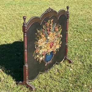 May include: An antique fireplace screen with a dark green background and a large floral arrangement in a blue vase. The screen has a decorative wooden frame with a dark brown finish and stands on two legs.
