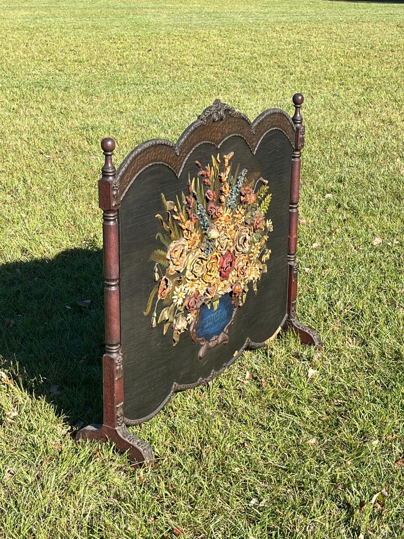 May include: An antique fireplace screen with a dark green background and a large floral arrangement in a blue vase. The screen has a decorative wooden frame with a dark brown finish and stands on two legs.