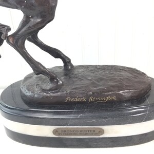 May include: Bronze sculpture of a horse bucking, titled "Bronco Buster by Frederic Remington". The sculpture is on a black marble base with a white marble band.