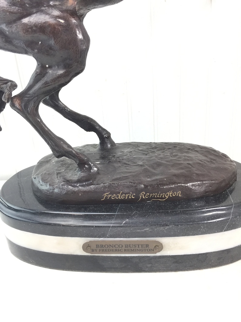 May include: Bronze sculpture of a horse bucking, titled "Bronco Buster by Frederic Remington". The sculpture is on a black marble base with a white marble band.