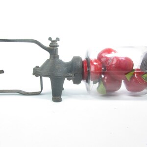 Puede incluir: Un molinillo de caf&eacute; vintage de hierro fundido con un frasco de vidrio lleno de manzanas rojas. El molinillo tiene una manija de madera y una manivela de metal.