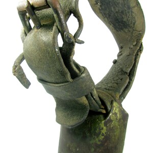 May include: Close-up of a weathered, green cowbell with a worn leather strap and a metal buckle. The strap is attached to the bell with a metal ring.
