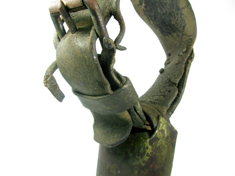 May include: Close-up of a weathered, green cowbell with a worn leather strap and a metal buckle. The strap is attached to the bell with a metal ring.