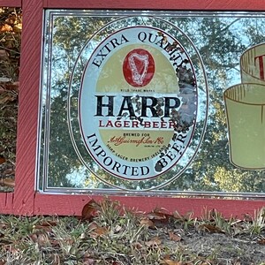 K&ouml;nnte beinhalten: Ein Vintage-Spiegel mit einem roten Holzrahmen. Der Spiegel spiegelt eine Harp Lager Beer-Werbung mit dem Text "Extra Quality", "Harp Lager Beer", "Brewed for Arthur Guinness Son & Co. Ltd.", "Harp Lager Brewery Ireland" und "Imported Beer" wider.