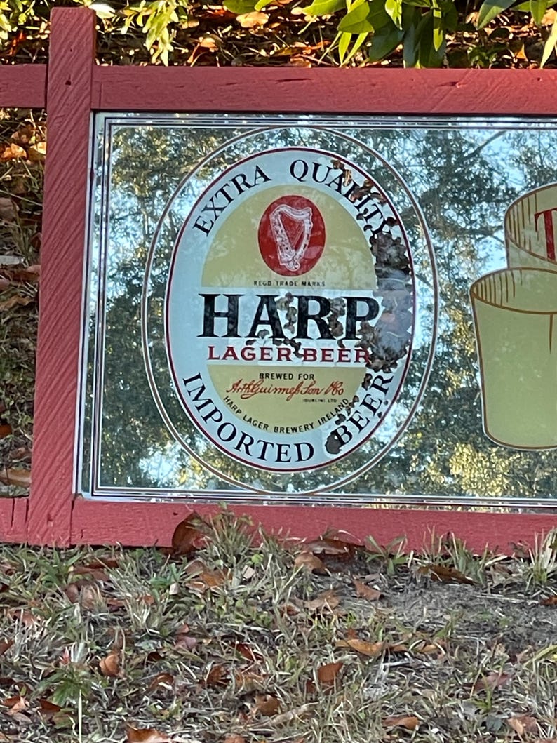 K&ouml;nnte beinhalten: Ein Vintage-Spiegel mit einem roten Holzrahmen. Der Spiegel spiegelt eine Harp Lager Beer-Werbung mit dem Text "Extra Quality", "Harp Lager Beer", "Brewed for Arthur Guinness Son & Co. Ltd.", "Harp Lager Brewery Ireland" und "Imported Beer" wider.