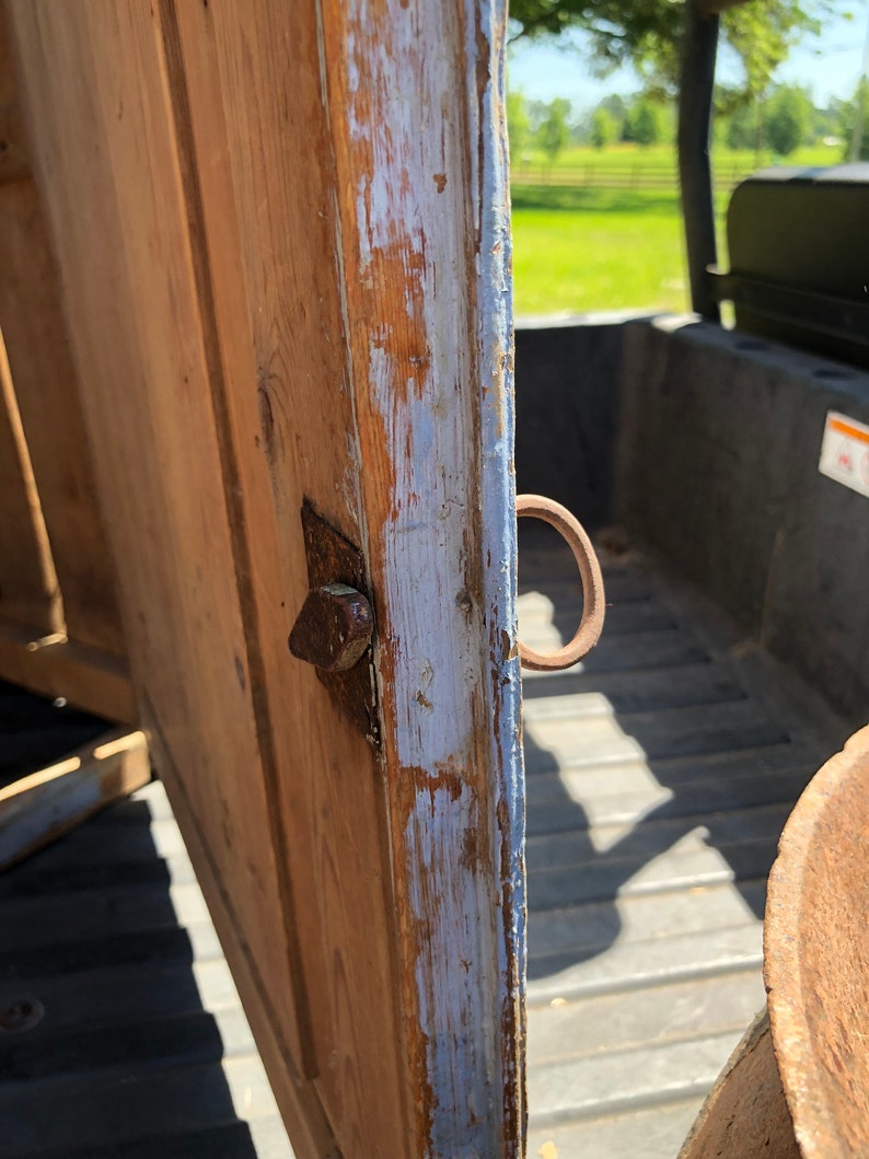 May include: Close-up of a wooden cabinet door with a metal hinge and a metal ring handle. The door is painted a light blue color and has some chipped paint. The wood is a light brown color.
