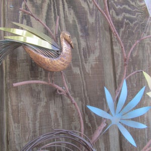 K&ouml;nnte beinhalten: Eine Wandskulptur mit einem stilisierten Vogel mit Kupfer- und Goldakzenten, der auf einem Ast mit Metallbl&auml;ttern in Blaut&ouml;nen, Gelb und Rosa sitzt.