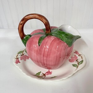 May include: A pink ceramic pitcher shaped like a peach with a brown handle and green leaves. The pitcher is sitting on a white ceramic plate with a floral design.