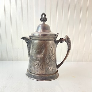 May include: A silver pitcher with an ornate design and a floral pattern. The pitcher has a lid with a decorative finial.