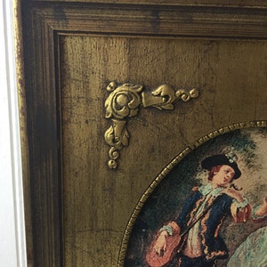 May include: An ornate gold-toned frame with a painting of a man in a blue jacket and a woman in a pink dress. The frame has a decorative corner piece.