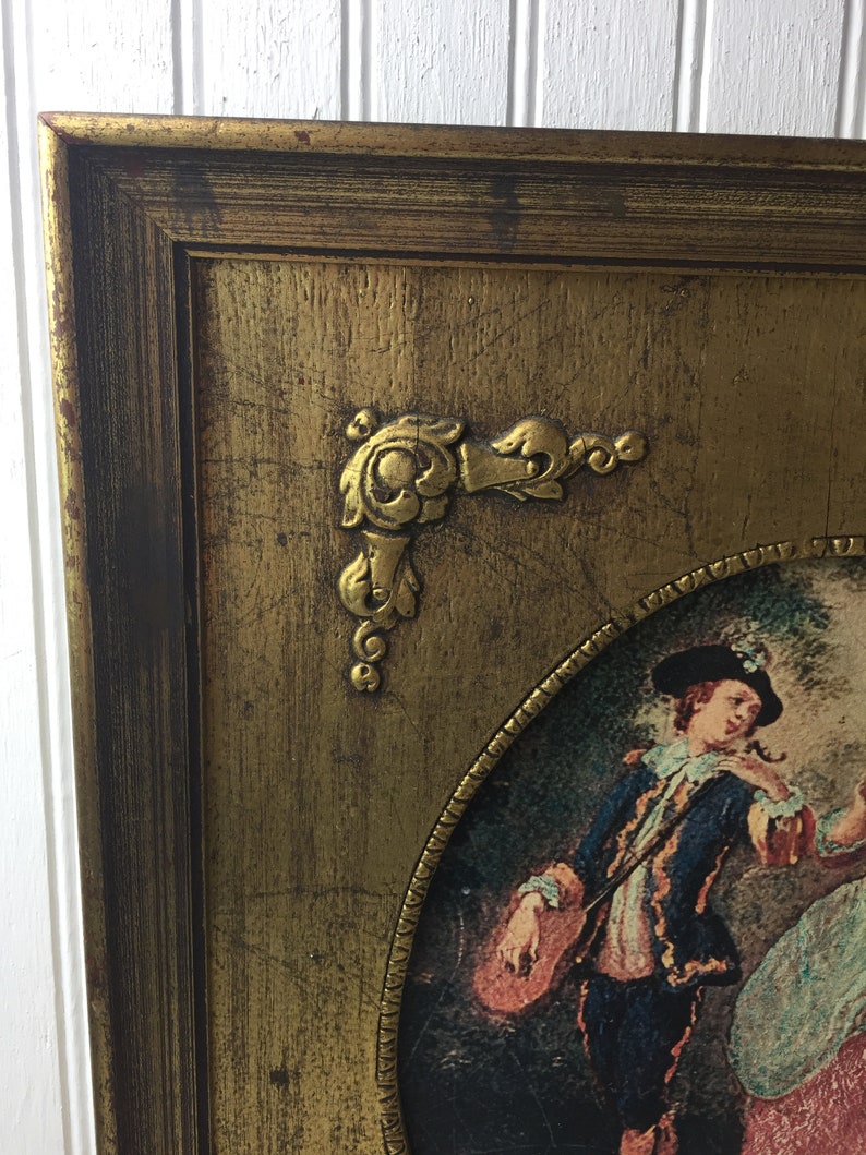 May include: An ornate gold-toned frame with a painting of a man in a blue jacket and a woman in a pink dress. The frame has a decorative corner piece.