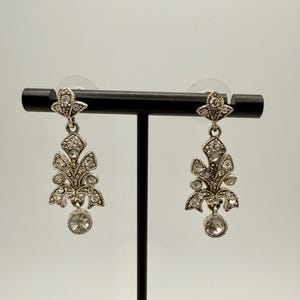 Op de afbeelding: Zilverkleurige oorhangers met een bloemmotief. Elke oorbel heeft bladvormige elementen versierd met kleine, heldere kristallen. Een grotere, ronde kristal hangt aan de onderkant van elke oorbel, wat glans toevoegt. De oorbellen worden op een zwarte standaard getoond.