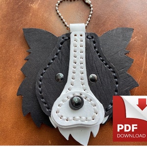 May include: A black and white leather keychain shaped like a dog's face. The keychain has a white nose and mouth with black eyes and a black chain attached to the top.