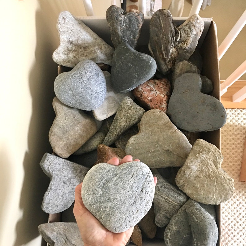 May include: A box filled with various heart-shaped stones in shades of gray, brown, and white. The stones are arranged in a random pattern, creating a visually appealing display.