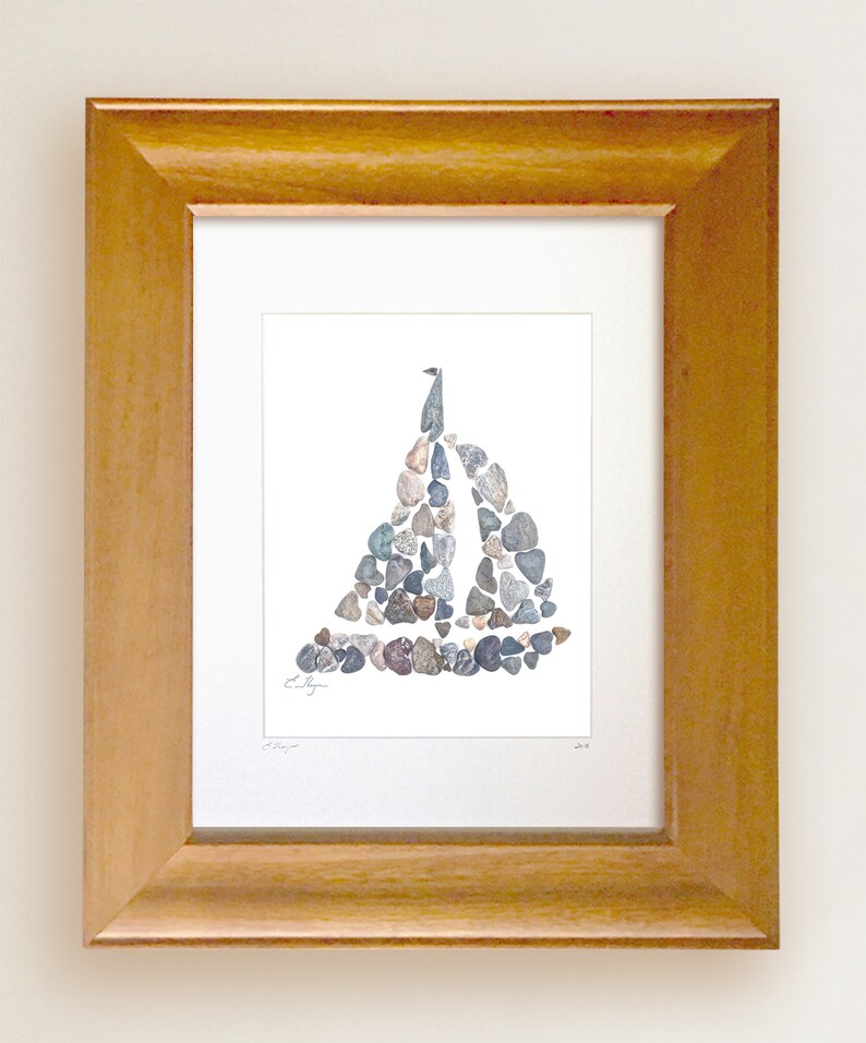 May include: A framed print of a sailboat made from small stones. The sailboat is a light gray color and is set against a white background. The frame is a light brown wood.