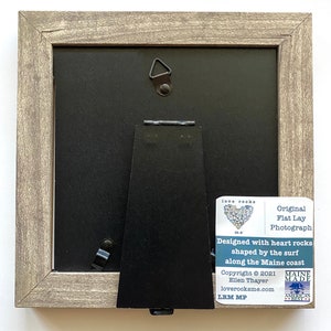 May include: A framed photograph of a heart made of flat, heart-shaped rocks. The frame is made of wood and has a gray finish. The back of the frame has a stand and a hanging hook. The photograph is titled "love rocks me" and is signed "LRM MP".