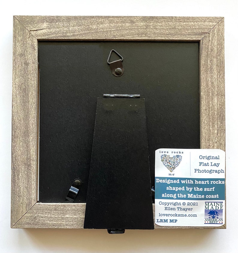 May include: A framed photograph of a heart made of flat, heart-shaped rocks. The frame is made of wood and has a gray finish. The back of the frame has a stand and a hanging hook. The photograph is titled "love rocks me" and is signed "LRM MP".