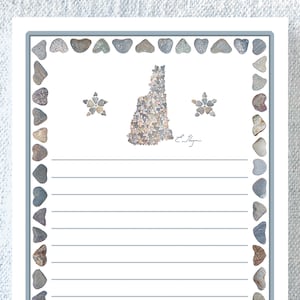 May include: A lined notepad with a white background and a border of gray stones. The state of New Hampshire is depicted in the center, made of small stones. Two star-shaped designs are also made of stones.