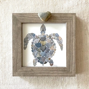 May include: A framed artwork featuring a sea turtle made from small, gray and brown stones. The turtle is made up of many heart-shaped stones. The frame is a light brown wood.