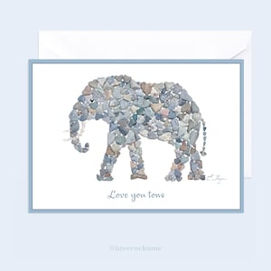 May include: A greeting card with a blue border features a white elephant made of small gray and brown heart-shaped stones. The text "Love you tons" is printed below the elephant.