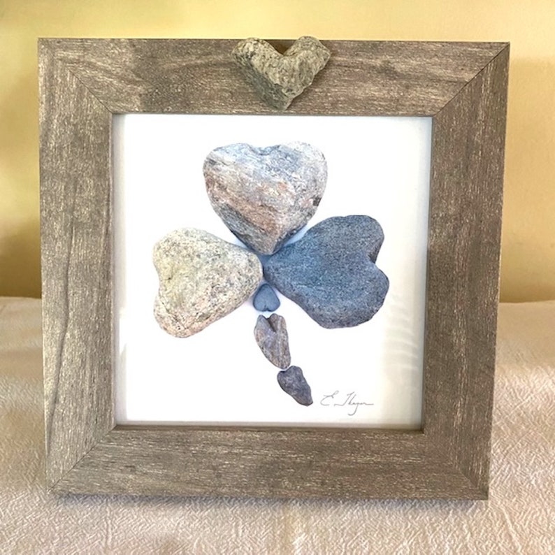 May include: A framed artwork featuring a shamrock made of gray and blue stones. The shamrock is surrounded by a white background and a wooden frame.