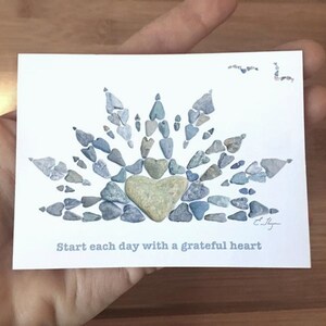 May include: A white card with a heart made of small blue and gray stones. The heart is surrounded by a sunburst design made of the same stones. The text "Start each day with a grateful heart" is printed below the heart.