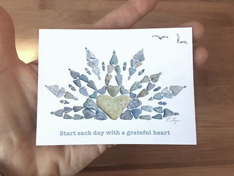 May include: A white card with a heart made of small blue and gray stones. The heart is surrounded by a sunburst design made of the same stones. The text "Start each day with a grateful heart" is printed below the heart.