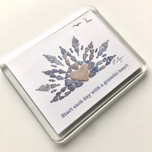 May include: A white card with a heart made of small gray and brown stones. The heart is surrounded by more stones in a sunburst pattern. The text "Start each day with a grateful heart" is printed on the card.