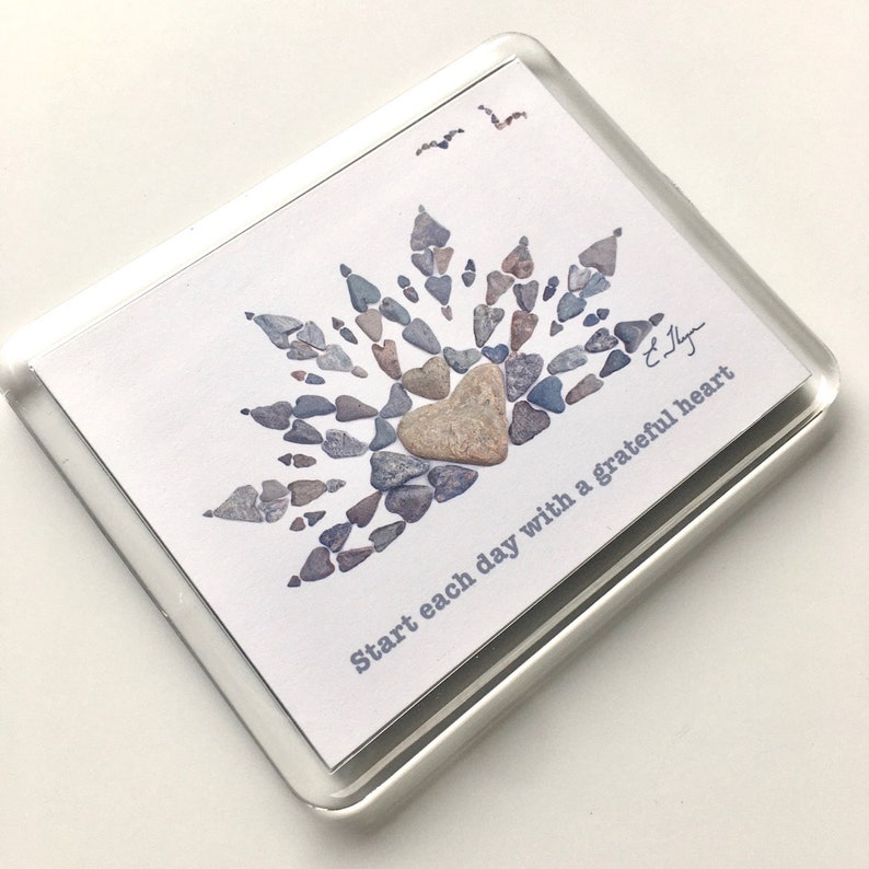 May include: A white card with a heart made of small gray and brown stones. The heart is surrounded by more stones in a sunburst pattern. The text "Start each day with a grateful heart" is printed on the card.