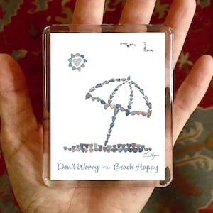 May include: A white background with a blue beach umbrella and a yellow sun made of small blue and gray heart-shaped stones. The text "Don't Worry ~ Beach Happy" is at the bottom of the image.