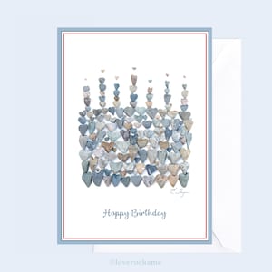 May include: A birthday card with a white background and a blue border. The card features a design of a cake made of small gray and blue heart-shaped stones. The text "Happy Birthday" is printed in black at the bottom of the card.